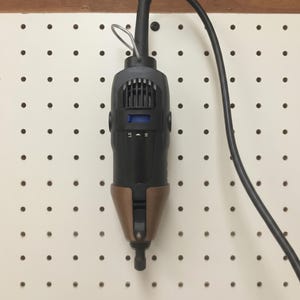 May include: A black and bronze power tool with a black power cord. The tool has a digital display and a bronze-colored lower section. It is hanging on a white pegboard.