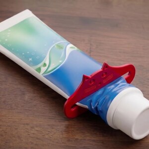 May include: A tube of toothpaste with a blue and white design, held by a red cat-shaped squeezer. The toothpaste tube is on a wooden surface.