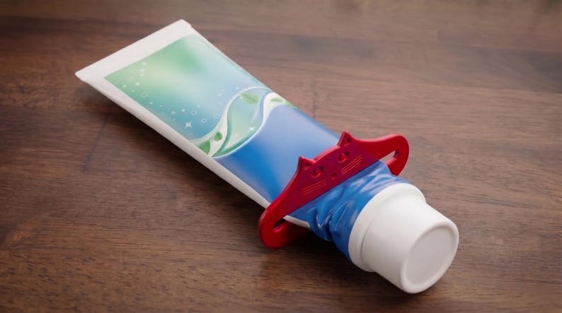 May include: A tube of toothpaste with a blue and white design, held by a red cat-shaped squeezer. The toothpaste tube is on a wooden surface.