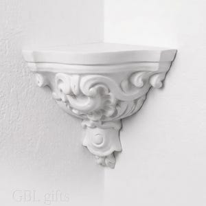 May include: A white corner wall shelf featuring ornate, carved scrollwork and floral-inspired detailing along its front edge. The shelf has a flat, triangular top surface and a decorative, tapered base. The text GBL gifts is visible at the bottom.