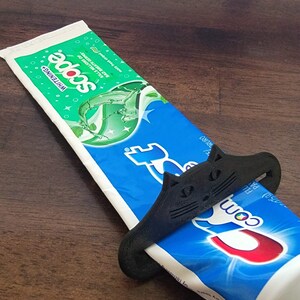 May include: A black cat-shaped toothpaste tube squeezer is wrapped around a blue and white tube of Crest toothpaste. The toothpaste tube is on a dark brown wooden surface. The toothpaste tube has the word "Crest" on it.
