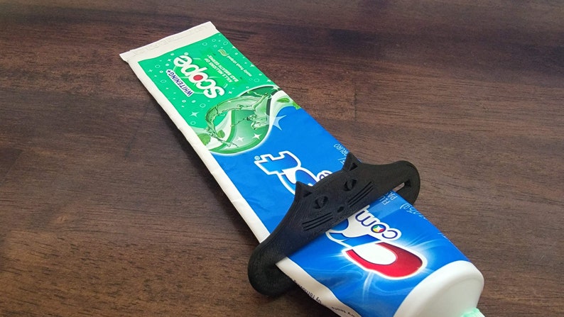 May include: A black cat-shaped toothpaste tube squeezer is wrapped around a blue and white tube of Crest toothpaste. The toothpaste tube is on a dark brown wooden surface. The toothpaste tube has the word "Crest" on it.