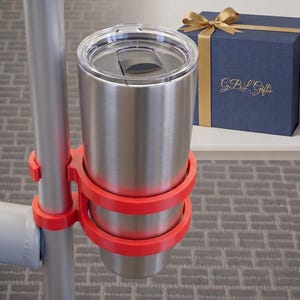 May include: A stainless steel tumbler sits in a red cup holder attached to a silver pole. A blue gift box with a gold ribbon and the text "GBL Gifts" is in the background, along with a small potted succulent.