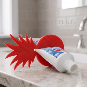 May include: A red plastic toothpaste tube squeezer in the shape of a pineapple. The squeezer is squeezing a tube of toothpaste. The product is on a bathroom countertop.