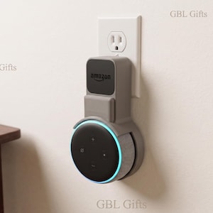 May include: A gray wall mount for an Amazon Echo Dot, plugged into a white electrical outlet. The Echo Dot is black with a blue light ring. The mount has the Amazon logo. A wooden table with a notebook, pen, and compass is in the background.