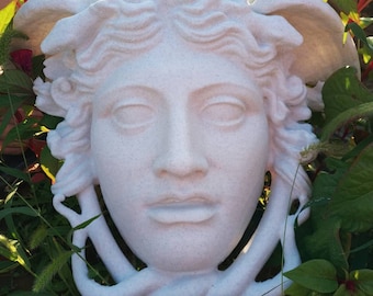 May include: A white, speckled, stone-like sculpture of a human face with wavy hair and winged features at the temples. The sculpture has a smooth, matte texture and is shaped with a rounded chin and defined facial features, including eyes and lips.
