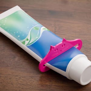 May include: A tube of toothpaste with a blue and green design, held by a pink cat-shaped squeezer. The tube is on a wooden surface.