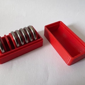 May include: A red 3D-printed ring holder with a rectangular shape, holding several silver rings. The holder is open, with a separate, empty red container next to it. The rings are evenly spaced.