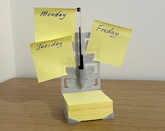 May include: A light gray desk organizer made of a textured material featuring a vertical back panel with a central pen holder and a base for a stack of square yellow sticky notes. Three yellow sticky notes are attached, labeled Monday, Tuesday, and Friday.
