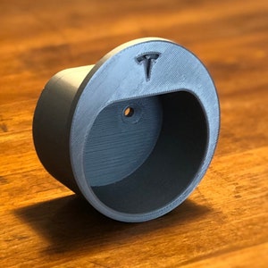May include: A gray, 3D-printed Tesla car charger holder. The holder is cylindrical with a curved opening and a small hole. The Tesla logo is visible on the top. The holder is made of a textured material.