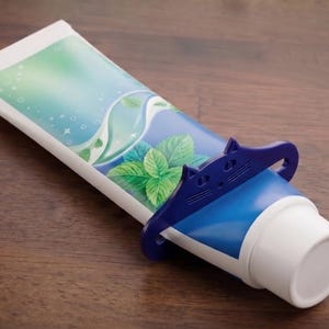 May include: A tube of toothpaste with a blue and white design, featuring a mint leaf graphic. A blue plastic tube squeezer is in use. The tube is on a wooden surface.