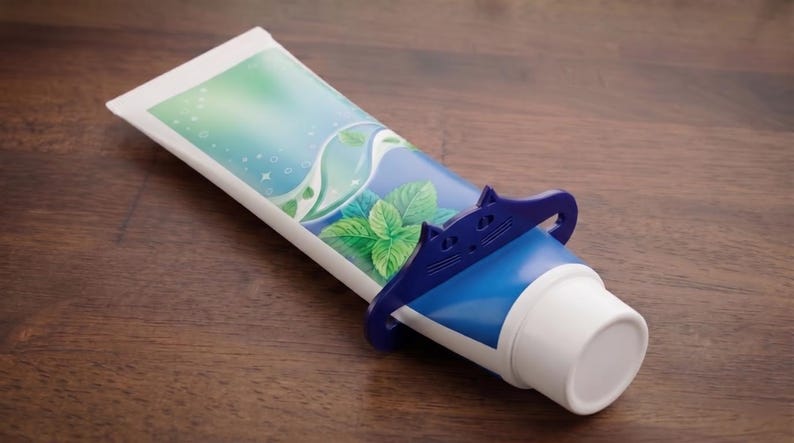 May include: A tube of toothpaste with a blue and white design, featuring a mint leaf graphic. A blue plastic tube squeezer is in use. The tube is on a wooden surface.
