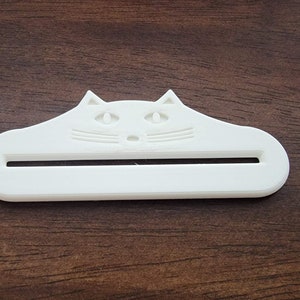 May include: A white cat-shaped object with a slot, likely a toothpaste tube squeezer. The cat's face is detailed with eyes, nose, and whiskers. The item is made of plastic and sits on a wooden surface.