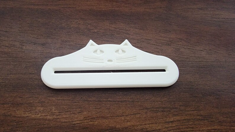 May include: A white cat-shaped object with a slot, likely a toothpaste tube squeezer. The cat's face is detailed with eyes, nose, and whiskers. The item is made of plastic and sits on a wooden surface.