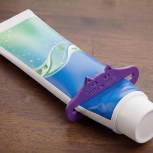 May include: A blue and white tube of toothpaste with a purple cat-shaped tube squeezer. The toothpaste tube has a blue and green design. The squeezer is made of plastic and is used to help get all the toothpaste out of the tube.