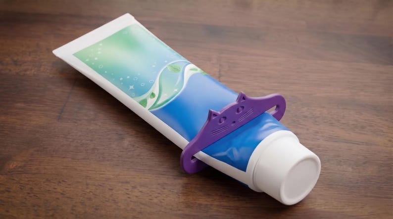 May include: A blue and white tube of toothpaste with a purple cat-shaped tube squeezer. The toothpaste tube has a blue and green design. The squeezer is made of plastic and is used to help get all the toothpaste out of the tube.