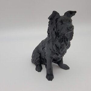 May include: A black 3D-printed dog figurine in a sitting position. The dog has a fluffy coat and detailed features, including perked ears and a focused gaze. The sculpture is set against a plain white background.