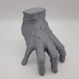 May include: A grey, three-dimensional, sculpted hand figurine featuring visible stitched seams across the wrist and knuckles. The hand is positioned in a downward-facing, walking stance with fingers curled, showcasing a textured, matte finish.