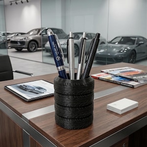 May include: A desk organizer made of stacked black tires holds pens. A blue pen with "LUXURY AUTO" printed on it is visible. The desk is made of wood and has a tablet, magazines, and business cards on it. Cars are visible in the background.