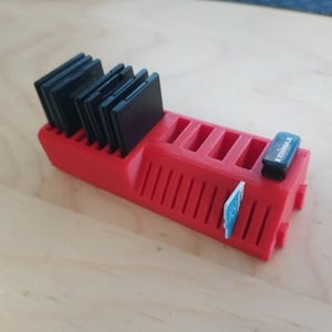 May include: A bright red 3D-printed storage rack featuring multiple vertical slots designed to hold several black SD memory cards. The unit includes a small black Edimax USB adapter and a single blue and white microSD card inserted into the side.