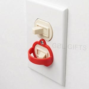 May include: A white electrical outlet cover with a light beige switch and a red switch guard. The switch guard has a loop design for easy operation. The text "GBL GIFTS" is visible on the cover.