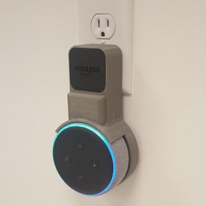 May include: A gray 3D printed wall mount for an Amazon Echo Dot smart speaker. The mount is attached to a standard electrical outlet. The Echo Dot is in the mount and has a blue light ring around it.