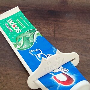 May include: A tube of Scope Whitening toothpaste being squeezed by a white cat-shaped toothpaste tube squeezer. The toothpaste tube is blue and green, and the squeezer is white. The background is a dark brown wooden surface.