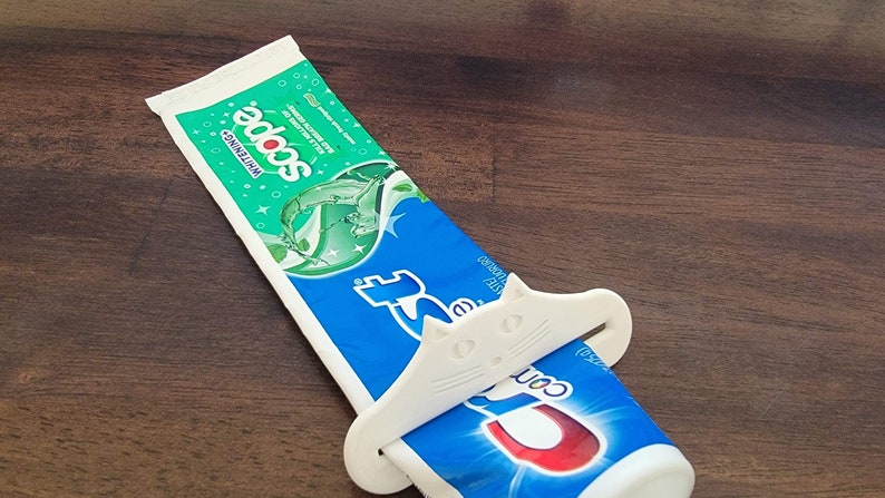 May include: A tube of Scope Whitening toothpaste being squeezed by a white cat-shaped toothpaste tube squeezer. The toothpaste tube is blue and green, and the squeezer is white. The background is a dark brown wooden surface.