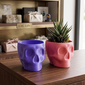 May include: Two skull-shaped planters, one purple and one pink, sit on a wooden surface. The pink planter holds a green and white striped succulent. The planters are next to each other. The text "GBL Gifts" is visible in the background.