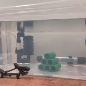 Pode incluir: Uma pequena figura de tubar&atilde;o de pl&aacute;stico preto fica em frente a um tanque de pl&aacute;stico transparente com um filtro verde, um filtro preto e um aquecedor. O tanque est&aacute; cheio de &aacute;gua.