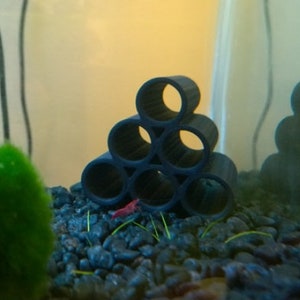 Pode incluir: Uma decora&ccedil;&atilde;o de aqu&aacute;rio de pl&aacute;stico preto em forma de pir&acirc;mide com seis aberturas redondas. A decora&ccedil;&atilde;o est&aacute; em um aqu&aacute;rio com bolas de musgo verde, cascalho preto e um camar&atilde;o vermelho.