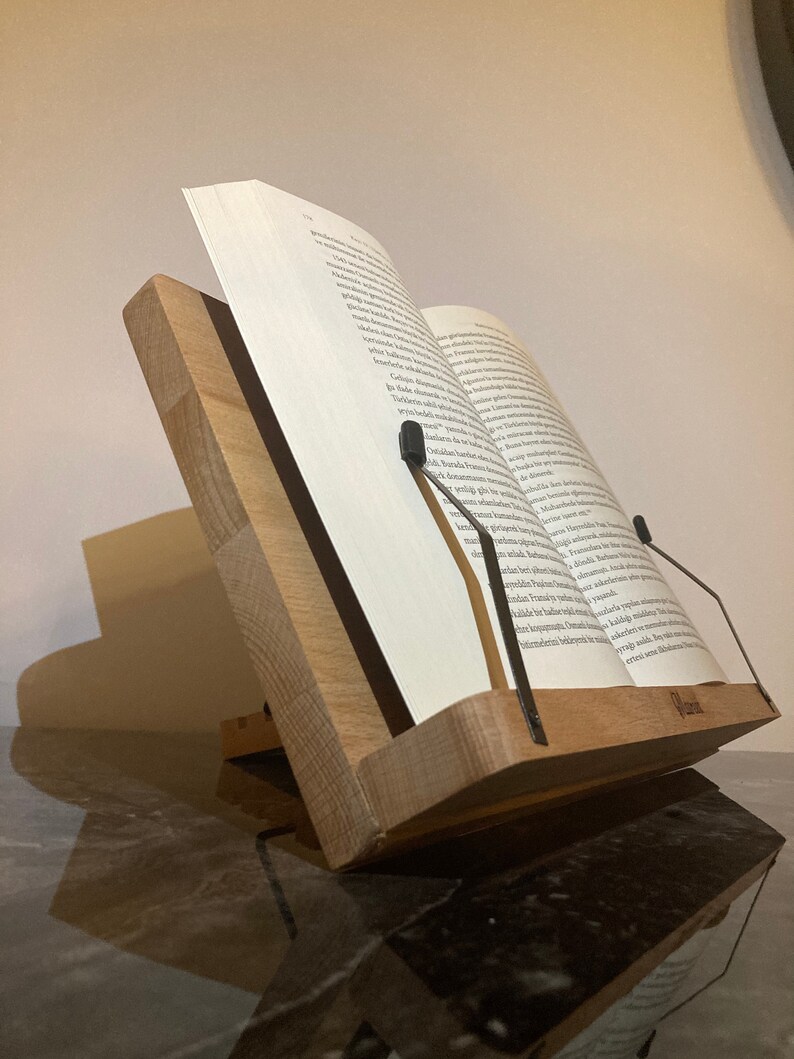 May include: A wooden book stand with metal page holders. The stand is made of natural wood and has a simple, elegant design. It is perfect for holding books, cookbooks, or other documents.