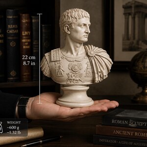 K&ouml;nnte beinhalten: Eine beige B&uuml;ste eines r&ouml;mischen Kaisers in verzierter R&uuml;stung mit detaillierten Reliefschnitzereien. Die Skulptur ist 22 cm hoch und wiegt etwa 600 g. Die Figur hat kurzes, strukturiertes Haar und einen &uuml;ber die Schulter drapierten Umhang.