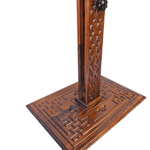 May include: A wooden stand with detailed geometric carvings. The stand has a rectangular base and a vertical support with a decorative pattern. A black knob is visible near the top. The wood has a warm, brown tone.