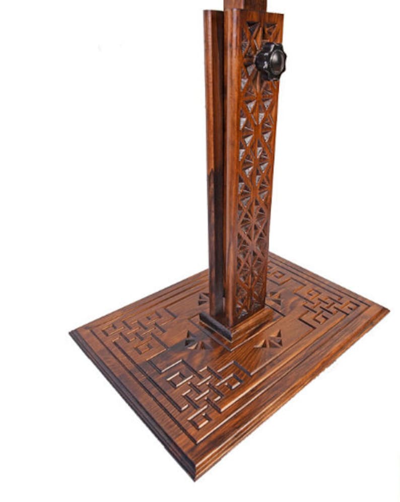 May include: A wooden stand with detailed geometric carvings. The stand has a rectangular base and a vertical support with a decorative pattern. A black knob is visible near the top. The wood has a warm, brown tone.