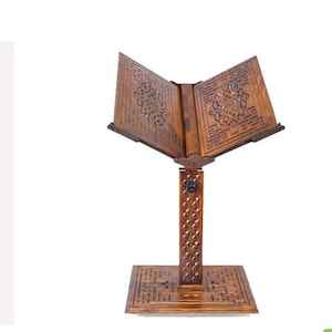 May include: A wooden book stand with detailed carved patterns. The stand is a warm brown colour and features a square base, a vertical support, and a book rest. The book rest is angled and has floral patterns.