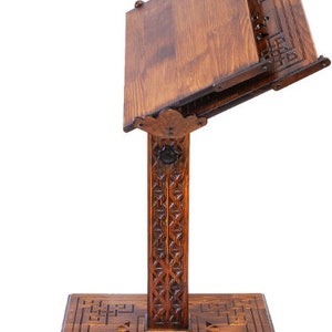 May include: A dark brown wooden book stand. It has a rectangular base and a vertical support with carved geometric patterns. The angled book rest has a decorative edge and is designed to hold books. The stand is made of wood.