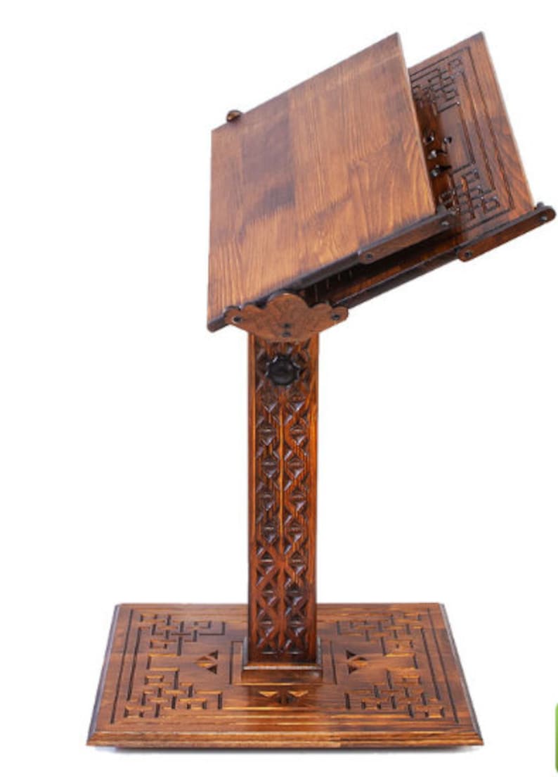 May include: A dark brown wooden book stand. It has a rectangular base and a vertical support with carved geometric patterns. The angled book rest has a decorative edge and is designed to hold books. The stand is made of wood.