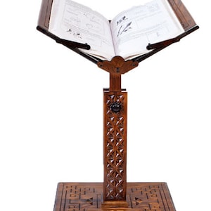 May include: A dark wood book stand with an open book. The stand has detailed carvings on the base and the vertical support. The book is open, showing text and illustrations. The stand is designed to hold books at an angle, ideal for reading.