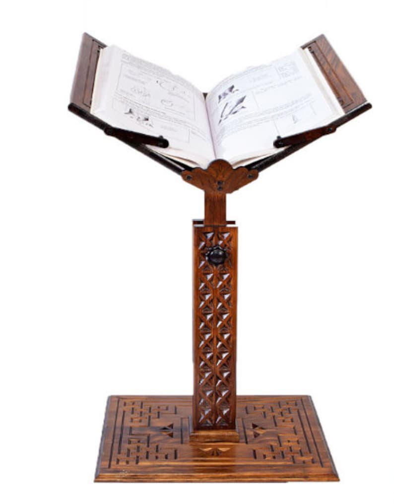 May include: A dark wood book stand with an open book. The stand has detailed carvings on the base and the vertical support. The book is open, showing text and illustrations. The stand is designed to hold books at an angle, ideal for reading.