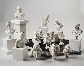 May include: A collection of white plaster busts of various figures, including men and women, arranged on a white surface with some on small white pedestals. The busts are all in a classical style, with detailed features and drapery.
