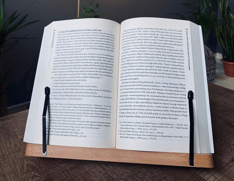 May include: A close-up of an open book with a black page holder. The book is open to a page with text in Turkish. The text discusses the history of the Faculty of Law at Istanbul University.