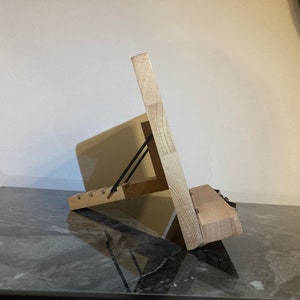 May include: A wooden stand with a black metal hinge, designed to hold a tablet or book at an angle. The stand is made of light-colored wood and has a natural finish.