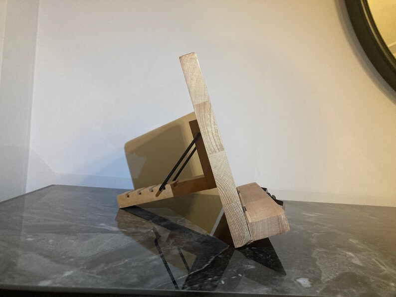 May include: A wooden stand with a black metal hinge, designed to hold a tablet or book at an angle. The stand is made of light-colored wood and has a natural finish.