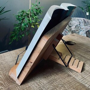 May include: A wooden book stand with a black metal page holder. The stand is made of natural wood and has a rustic look. The stand is designed to hold a book open at an angle for easy reading.