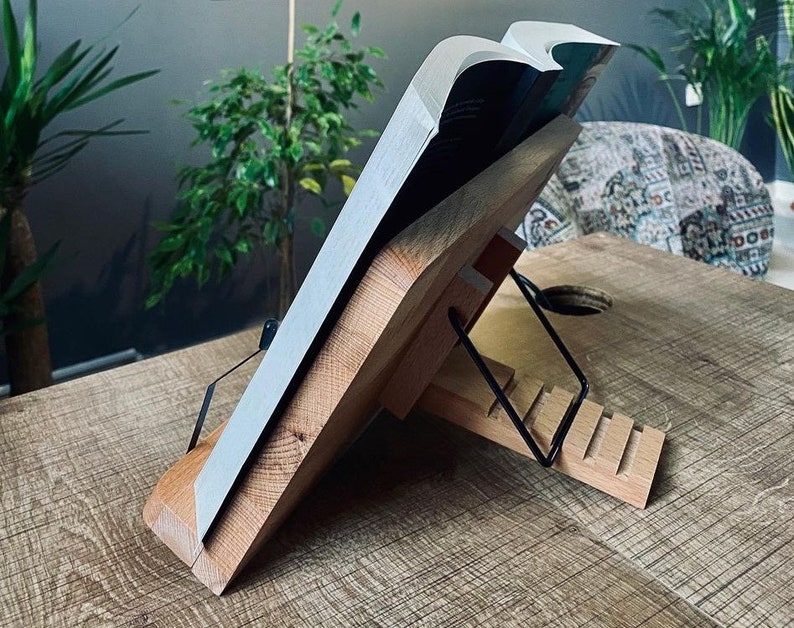 May include: A wooden book stand with a black metal page holder. The stand is made of natural wood and has a rustic look. The stand is designed to hold a book open at an angle for easy reading.