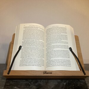 May include: A wooden book stand with two black page holders. The stand has the word "Maureen" engraved on the front. A book is resting on the stand, with the pages open.