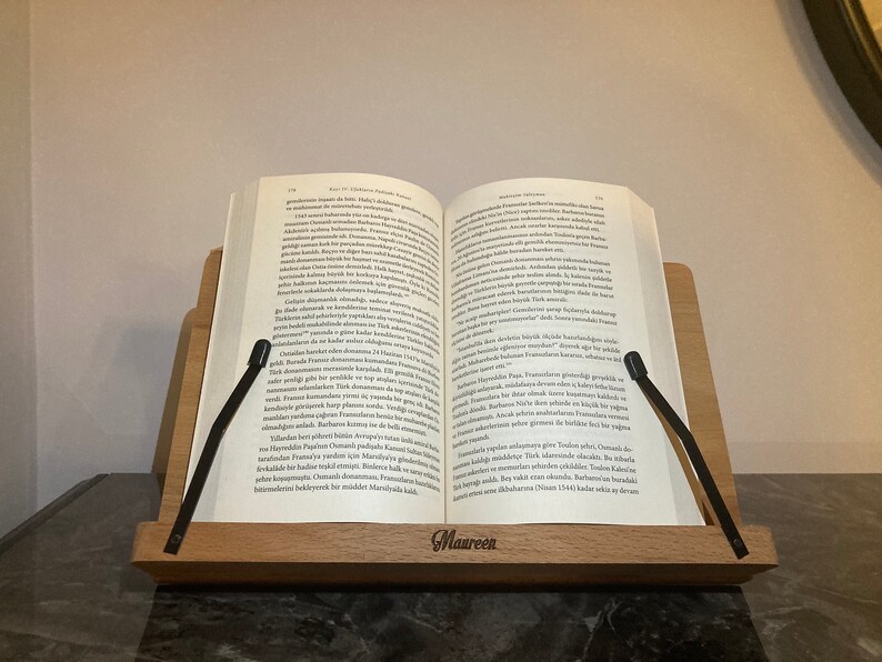 May include: A wooden book stand with two black page holders. The stand has the word "Maureen" engraved on the front. A book is resting on the stand, with the pages open.