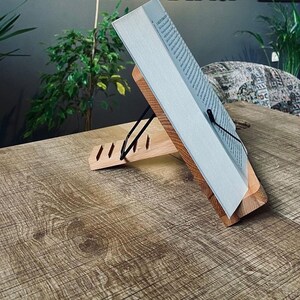 May include: A wooden book stand with a book resting on it. The stand has a black metal clip to hold the book in place. The stand is on a wooden table.