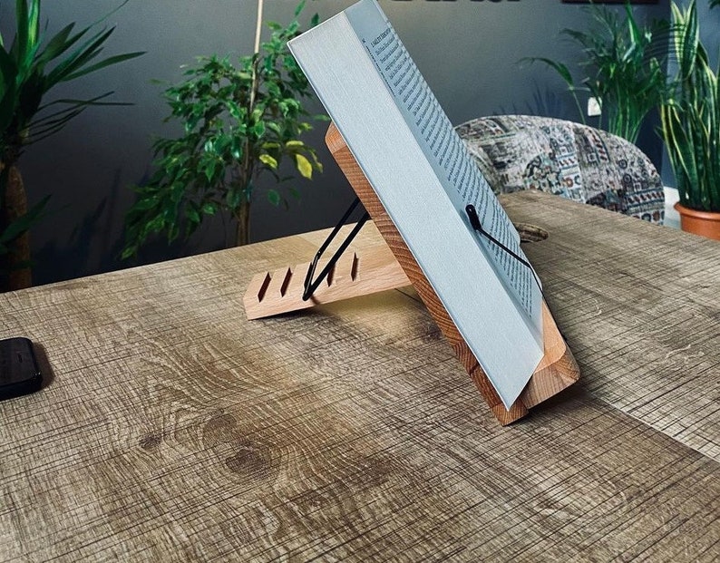 May include: A wooden book stand with a book resting on it. The stand has a black metal clip to hold the book in place. The stand is on a wooden table.